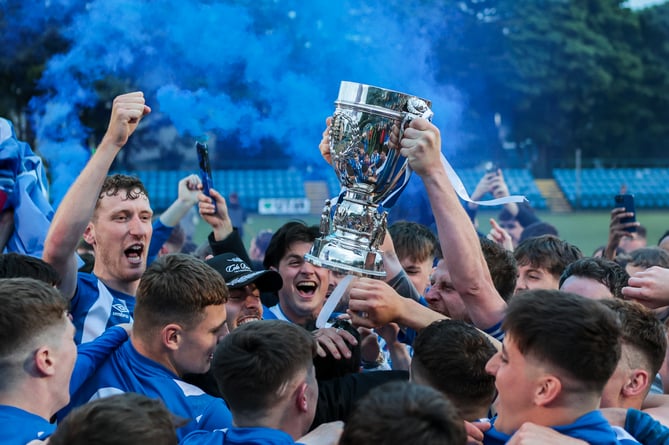 With blue smoke bombs going off in the background, Ramsey players celebrate with the Hospital Cup after Friday evening's final at the Bowl (Photo: Gary Weightman)